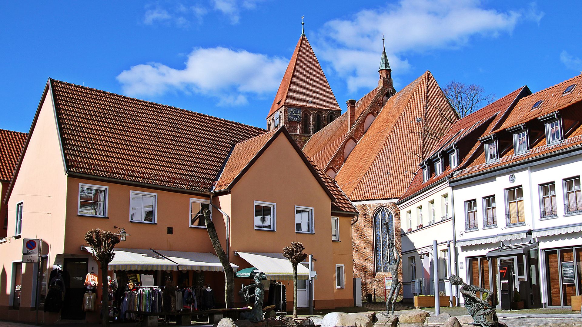 Blik auf Brunnen, ein paar Häuser und eine Kirche im Hintergrund vor blauem Himmel