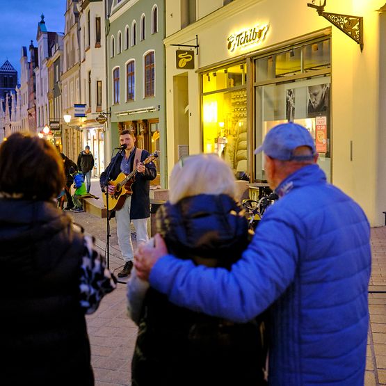 Der Sänger Öxl singt und spielt Gitarre in der Wismarer Fußgängerzone. Mehrere Personen schauen ihm zu.