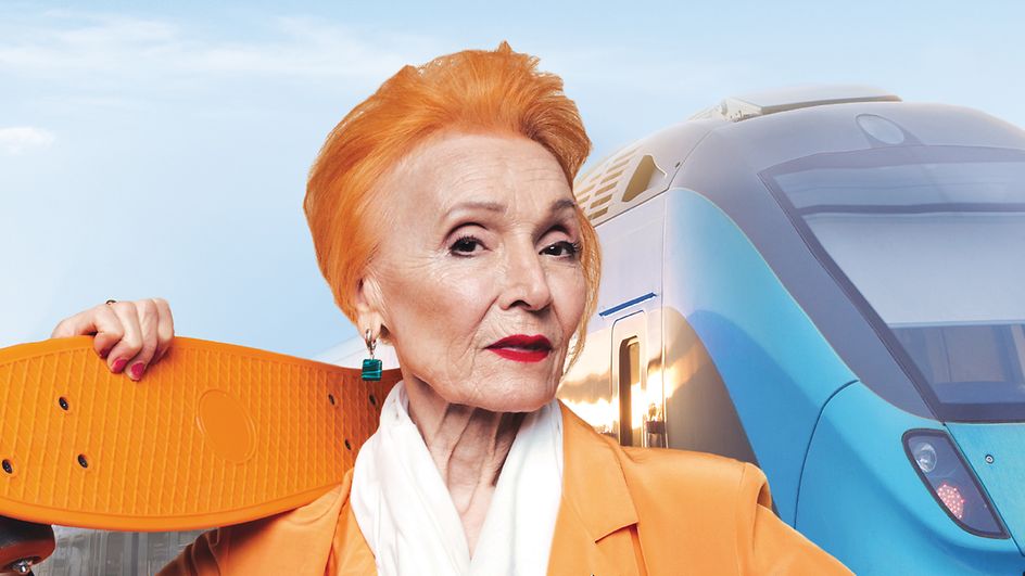 Eine ältere Dame mit orange gefärbtem Haar und geschultertem Penny-Board steht vor einem Zug