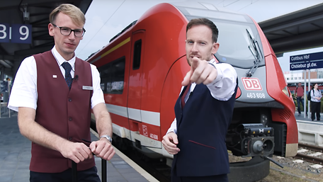Der Influence Benny Job steht mit einem Kundenbetreuer der DB vor einem roten Regionalzug.