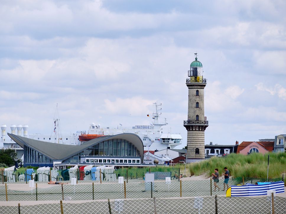 Strandkörbe vor einem Haus mit geschwungenem, dem Warnemünder Teepott, daneben der Leuchtturm, dahinter eine weiße Fähre