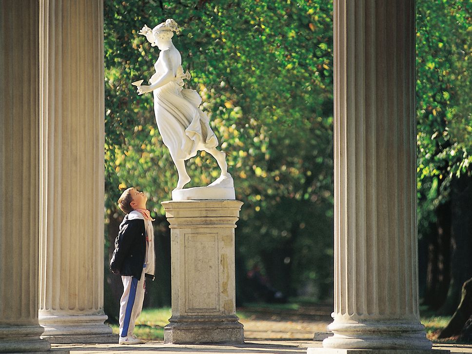 Ein Junge steht zwischen Säulen und schaut hinauf zur weißen Statue einer Frau auf einem Sockel im Schlosspark Neustrelitz