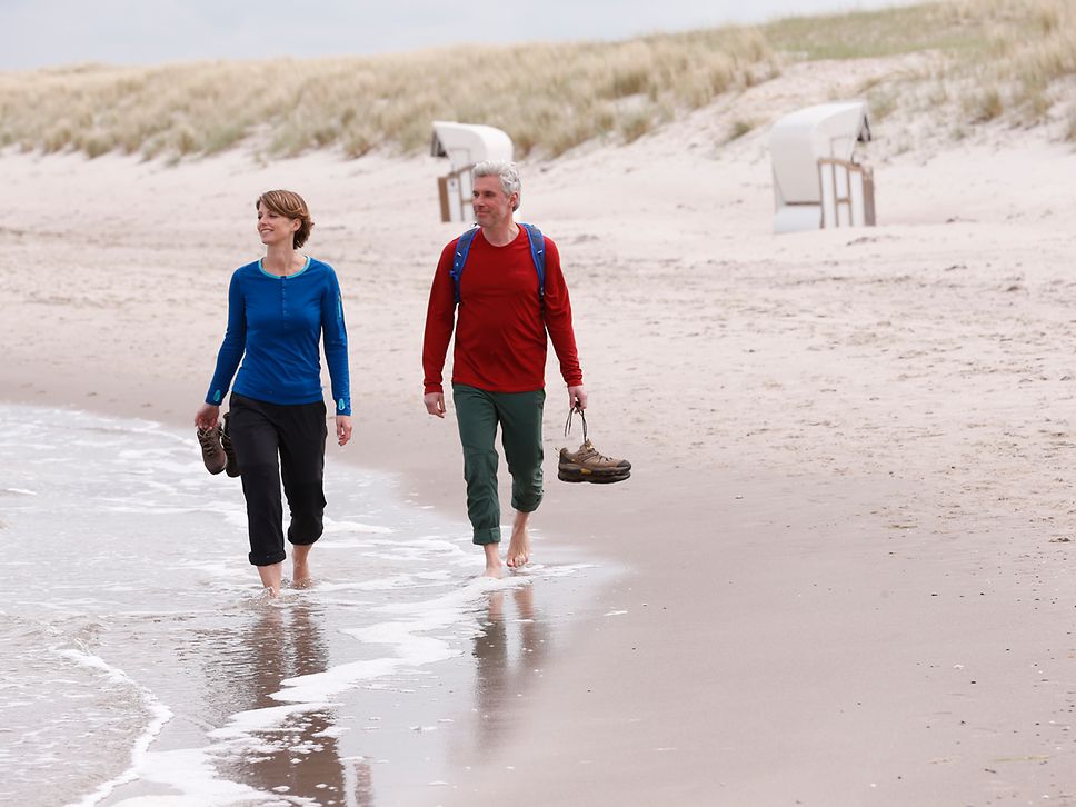 Eine Frau und ein Mann mit grauen Haaren barfuß im flachen Wasser am Strand, die Schuhe in der Hand, dahinter beginnen die Dünen