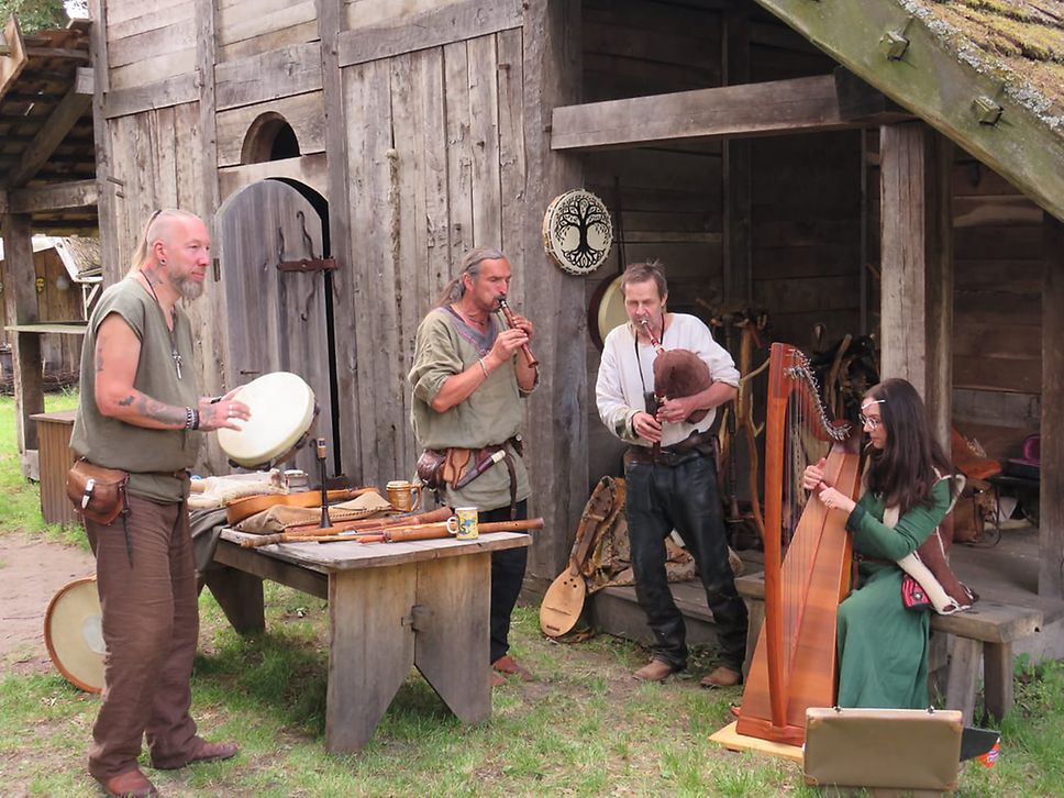 Drei Musiker spielen vor nachgebauten Holzgebäuden aus dem Mittelalter auf alten Instrumenten, eine Musikerin spielt Harfe