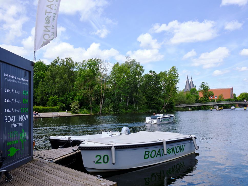 Hölzerner Bootssteg mit weißem Boot von Boat Now an der Obertrave in Lübeck, im Hintergrund die Türme des Holstentors