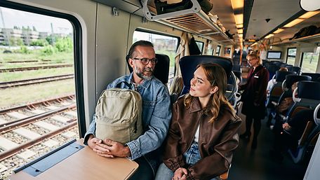 In einem Regionalzug: Mann mit Rucksack auf dem Schoß sitzt neben junger Frau. Die beiden schauen sich freundlich an.
