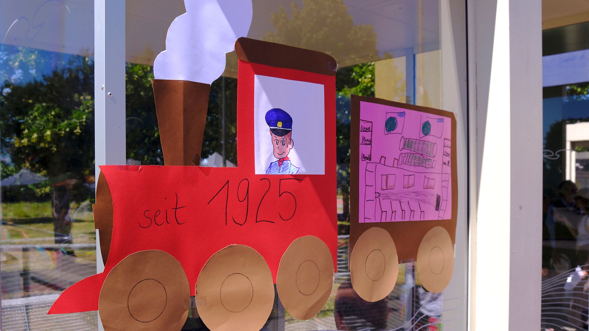 Ausschnitt von kindlich gestalteter Eisenbahn aus bunter Pappe mit der Aufschrift „seit 1925“ an Wartehäuschen auf dem Bahnsteig
