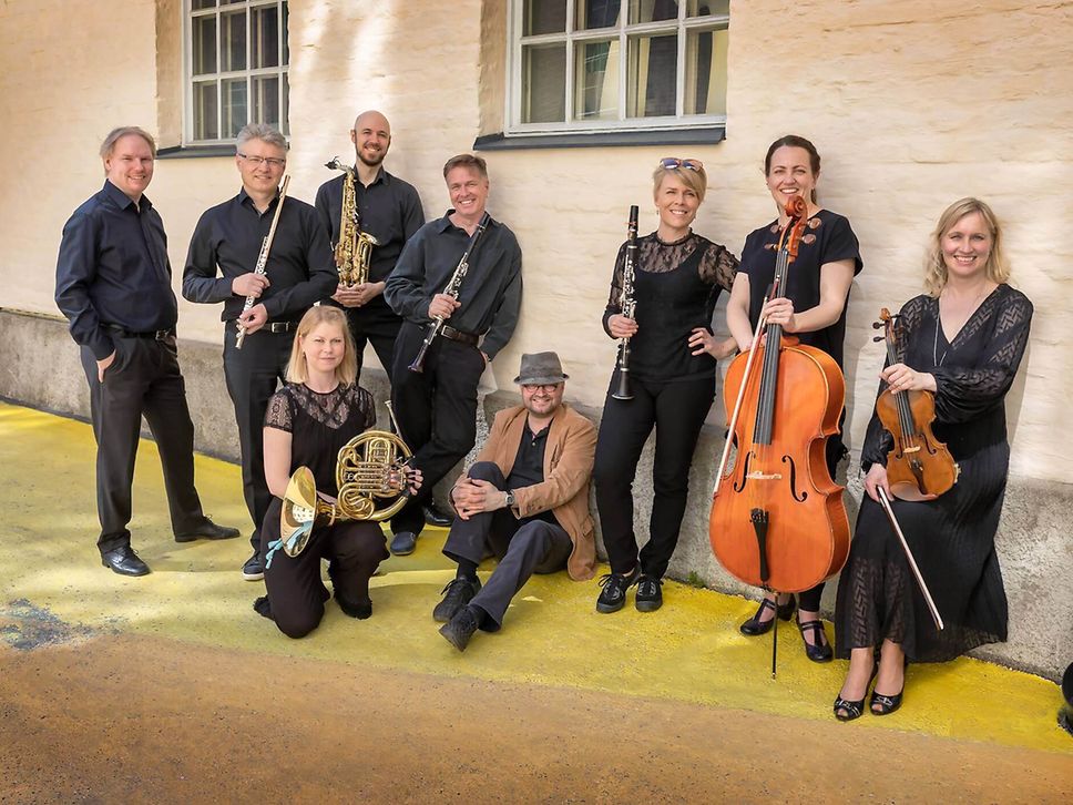 Neun Musikerinnen und Musiker des finnischen Ensembles TampereRaw posieren mit ihren Instrumenten vor einer Hauswand.