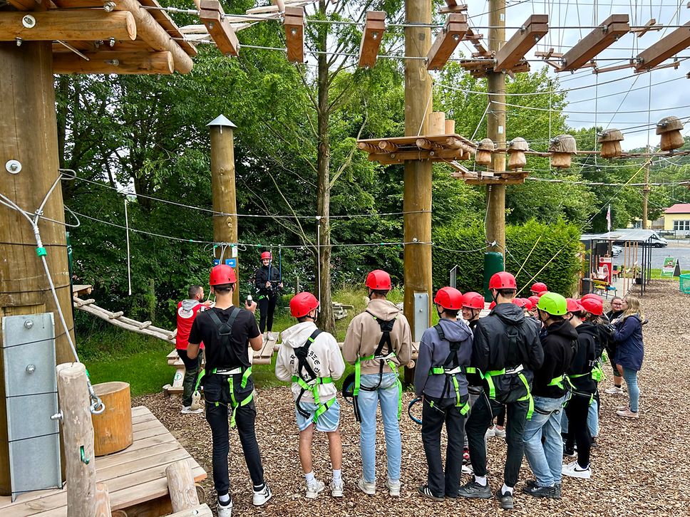 Eine Gruppe von Kletterern mit Helmen und Sicherungsgurten steht vor einem Teammitglied des Kletterparks in Burg Stargard