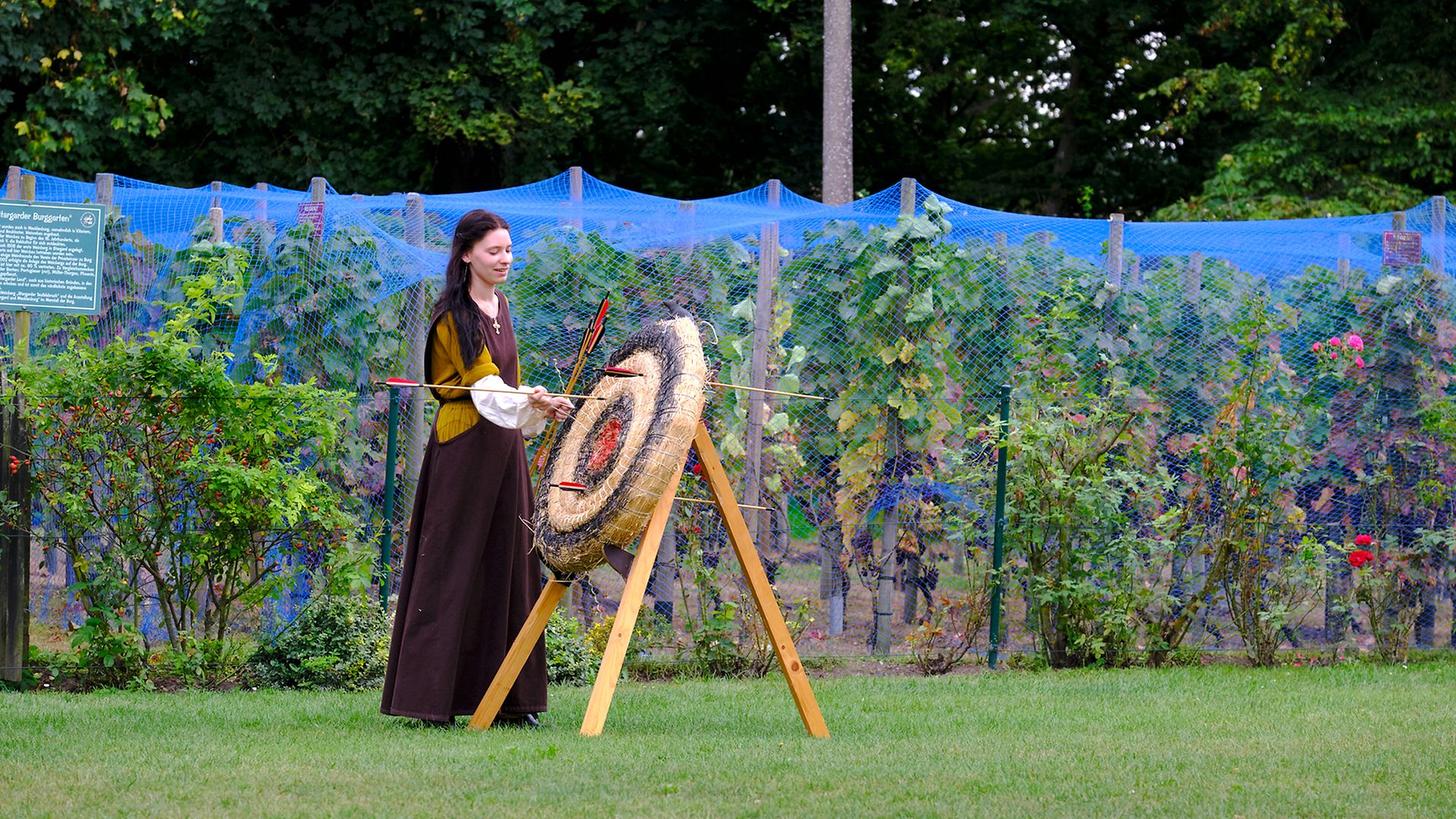Lilith Köhler in mittelalterlichem Kostüm und mit Pfeil und Bogen neben einer Zielscheibe auf Burg Stargard