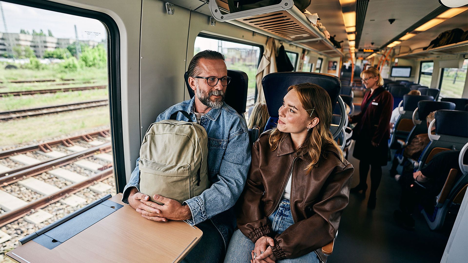 Ein Mann mit Rucksack auf dem Schoß sitzt in einem Viererabteil neben einer Frau, sie unterhalten sich.