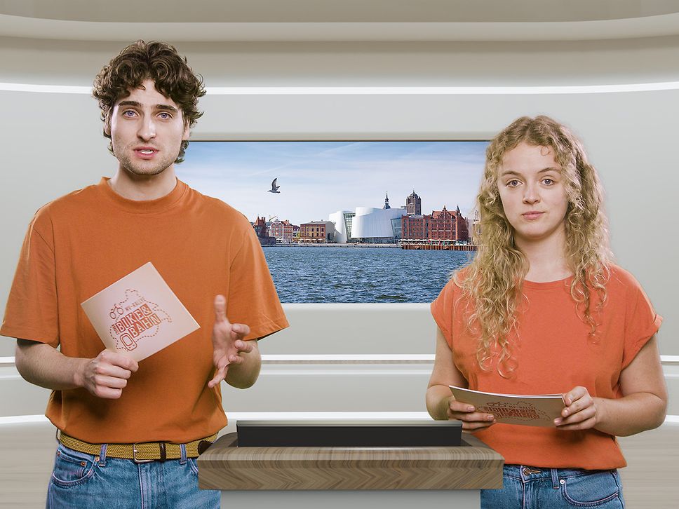 Zwei Studierende stehen in einem Studio und halten Karteikarten in den Händen, im Hintergrund ein Bild von Stralsund 