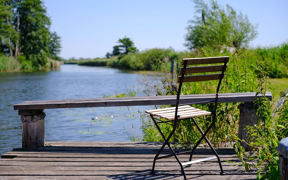 Blick von einem Holzsteg auf den Fluss Peene, im Vordergrund ein Gartenstuhl aus Holz.