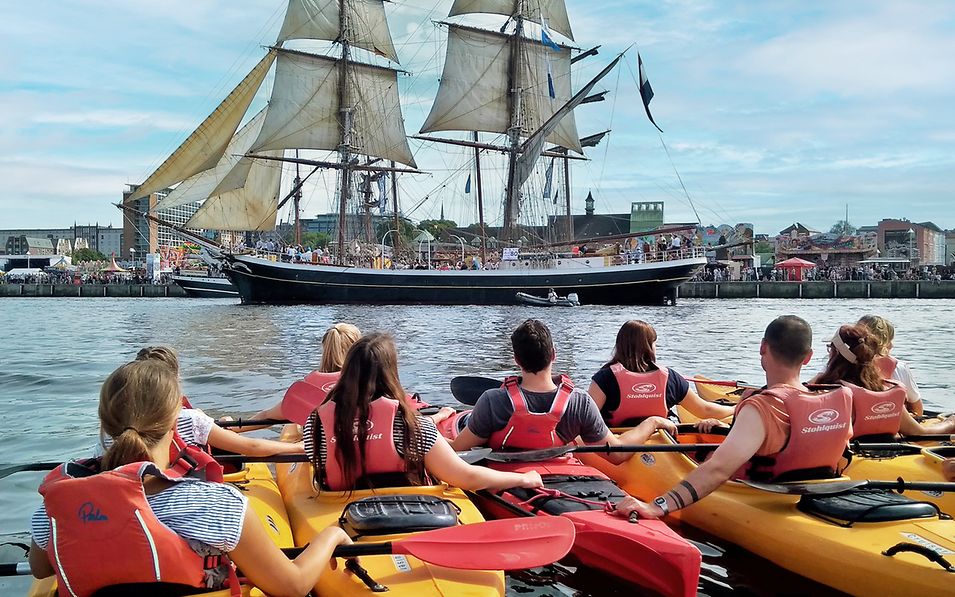 Schüler:innen in gelben Kajaks schauen auf ein Segelschiff vor der Stadtsilhouette von Rostock 