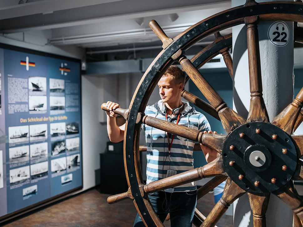 Ein junger Mann steht an einem mehr als mannshohen Steuerrad im Schifffahrtsmuseum Rostock