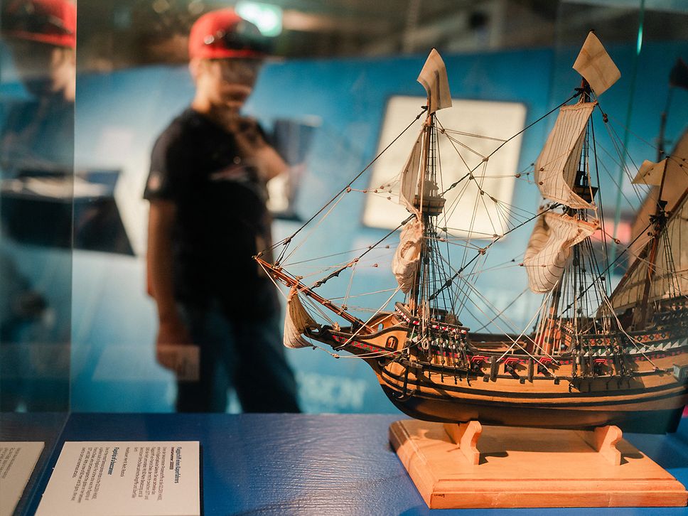 Ein Kind mit rotem Käppi steht vor einer Vitrine mit einem Schiffsmodell im Schifffahrtsmuseum Rostock