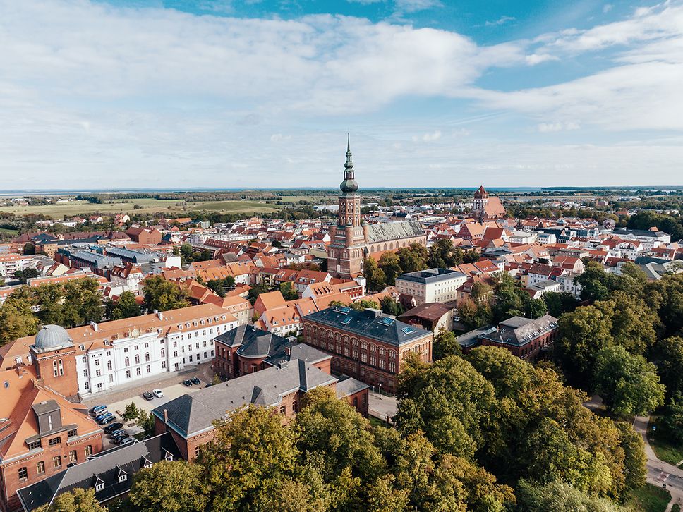 Die weißen und roten Häuser der Greifswalder Innenstadt rund um den Dom, eingebettet in die grüne Umgebung