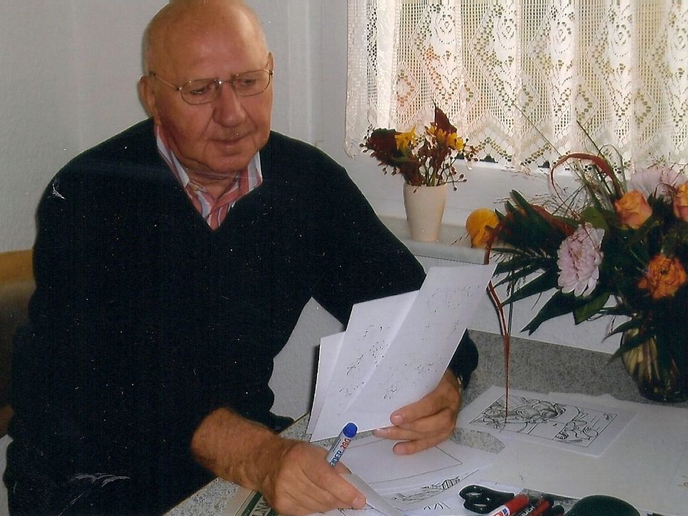 Der Karikaturist Günter Endlich an einem Tisch am Fenster, in der Hand mehrere Zettel mit eigenen Zeichnungen