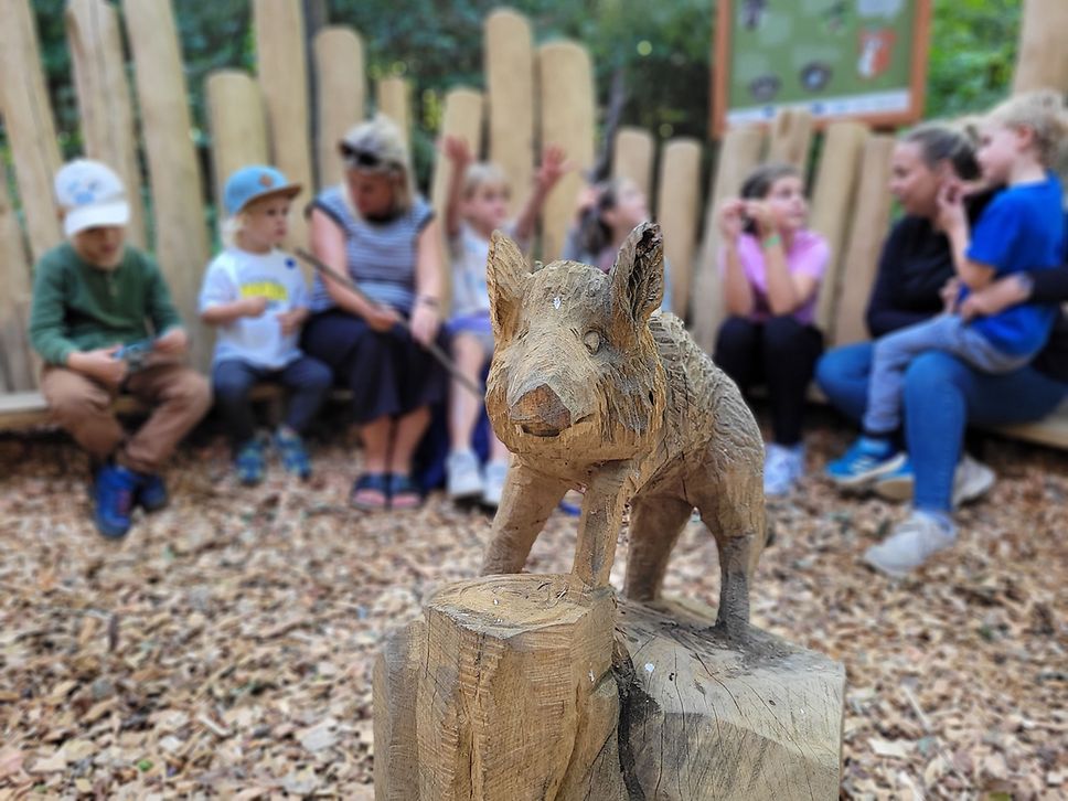 Mehrere Kinder sitzen auf Holzbänken rund um die Holzskulptur eines Wildschweins