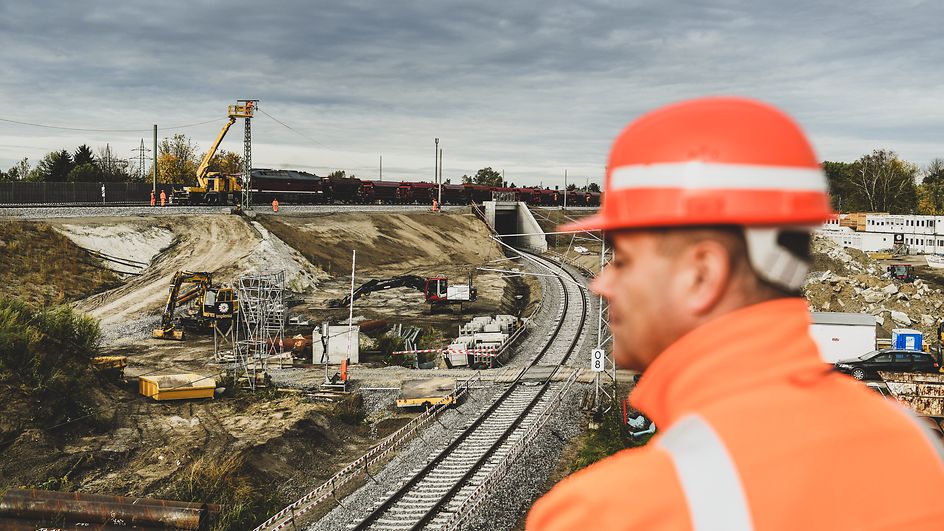 Bauarbeiter steht mit oranger Jacke und Helm etwas erhöht. Im Hintergrund ist eine Baustelle an einer Bahnstrecke zu sehen