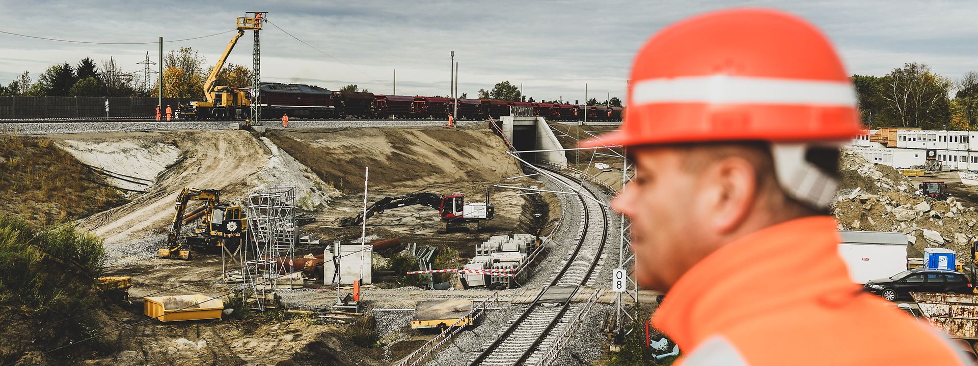 Bauarbeiter steht mit oranger Jacke und Helm etwas erhöht. Im Hintergrund ist eine Baustelle an einer Bahnstrecke zu sehen