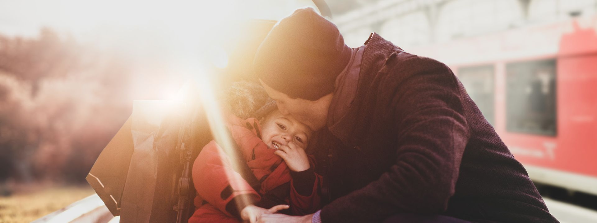 Quelle: istock.com / AleksandarNakic Ein Vater hockt sich zu seinem Kind, das im Buggy sitzt und gibt dem Kind einen Kuss auf die Stirn. Sie warten am Gleis.