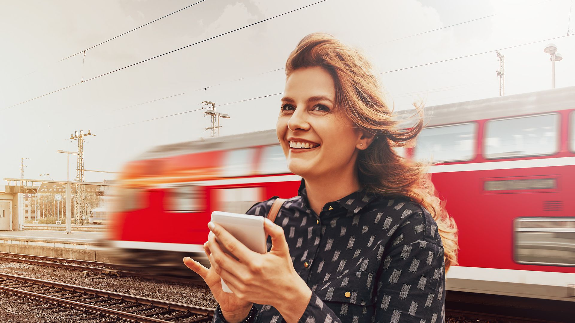Frau steht mit Handy in der Hand zufrieden am Bahngleis. Im Hintergrund fährt eine Regionalbahn ein.