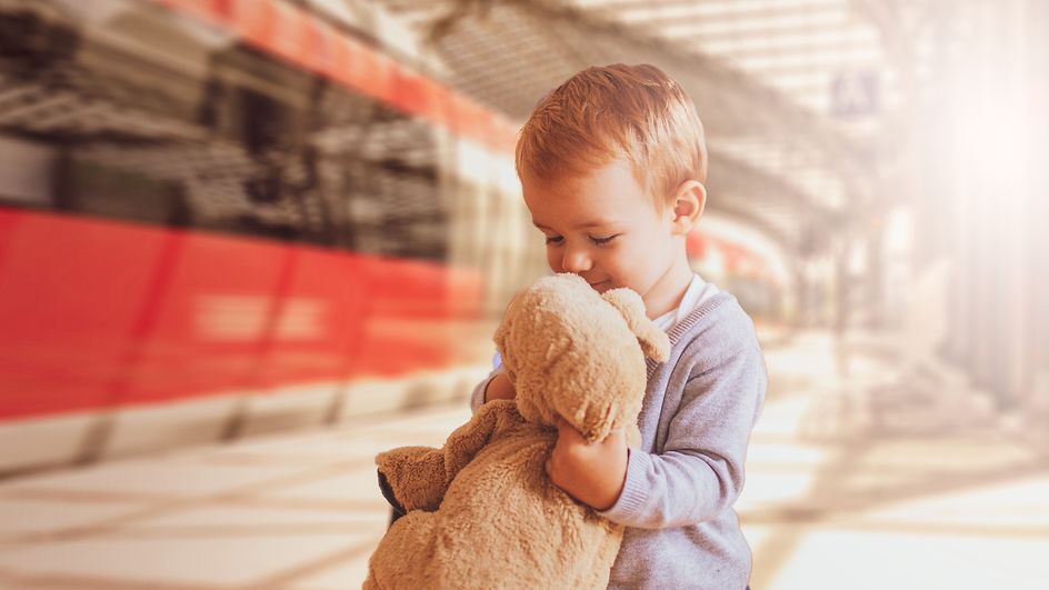 Kleiner Junge steht am Bahnhof und umarmt glücklich seinen Teddy, den er wiedergefunden hat. 